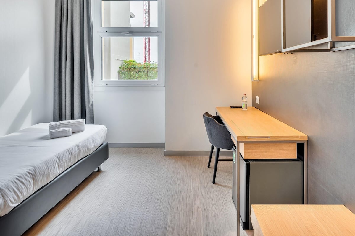 Ein modernes Schlafzimmer mit Bett, Schreibtisch und Fenster mit Blick auf ein Gebäude und Grünflächen.