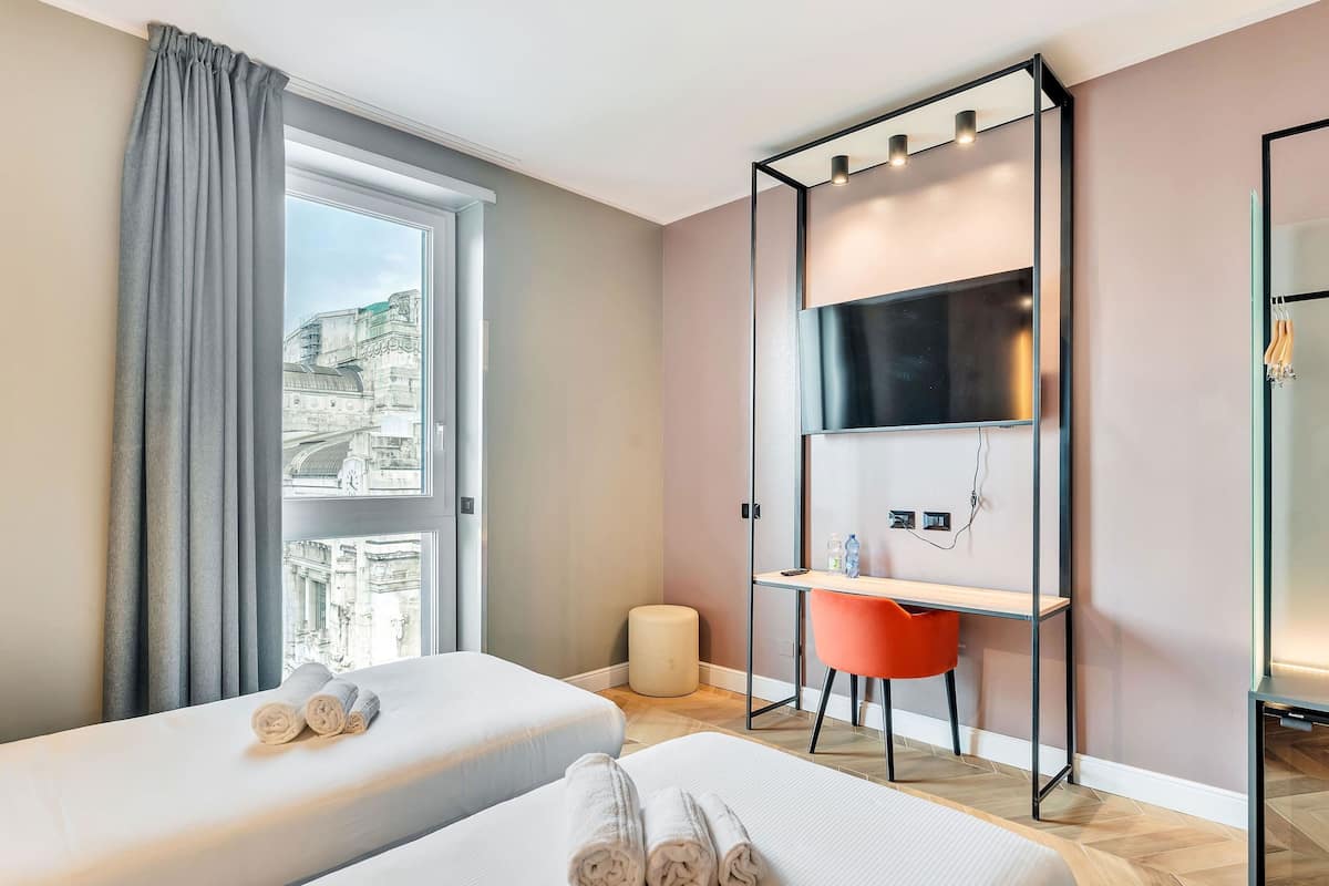 Ein Hotelzimmer mit zwei Betten, einem Schreibtisch, einem Stuhl, einem Fernseher und einem Fenster mit Vorhängen.