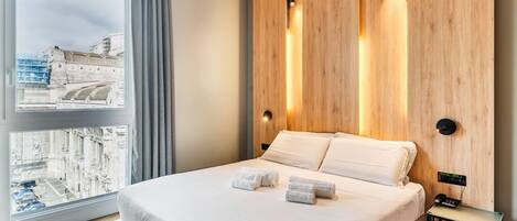 A modern hotel room with a large bed, a view of a cityscape, wooden paneling, and a bedside table with a phone.