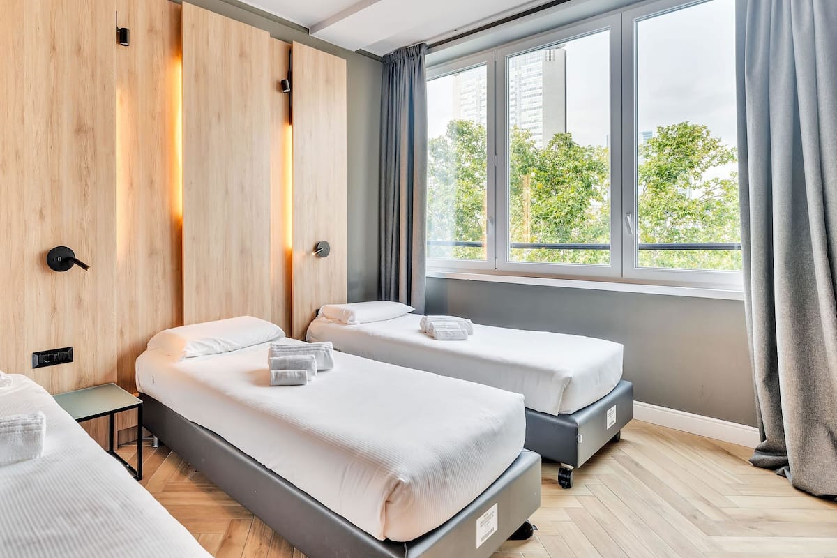 Ein Hotelzimmer mit zwei Betten, einem Holzschrank, einem Fenster mit Vorhängen und Blick auf Grünfläche.