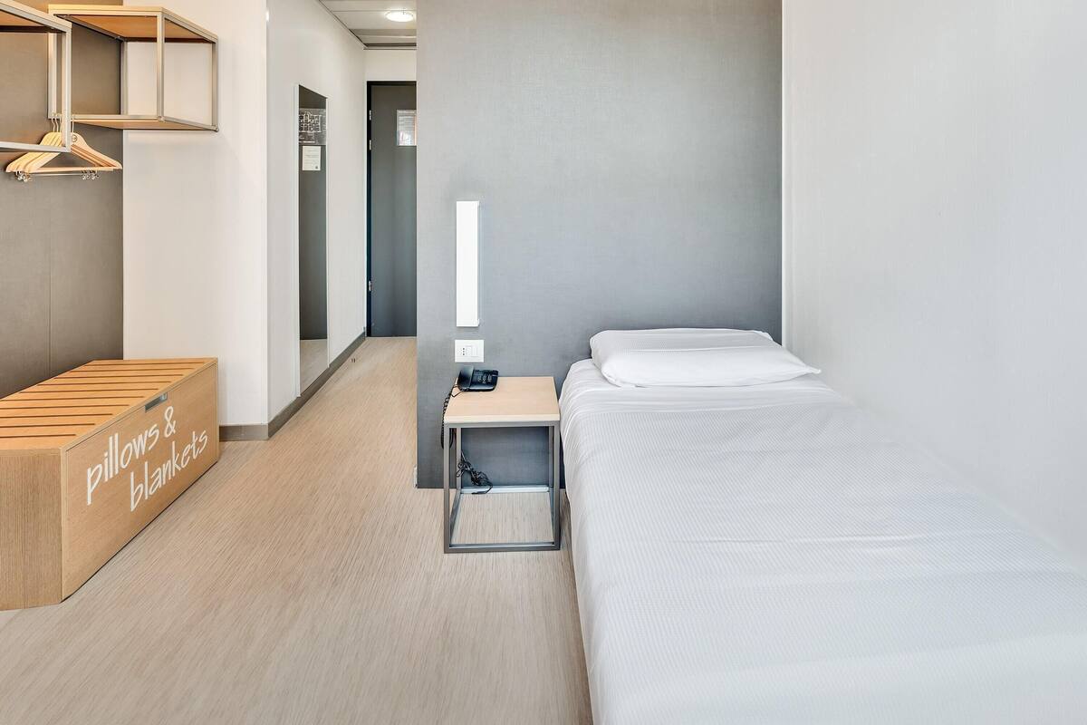 Ein Hotelzimmer mit einem Bett, einem Nachttisch mit Telefon, einer Holzkiste mit der Aufschrift 'pillows & blankets' und einer Pendelleuchte.