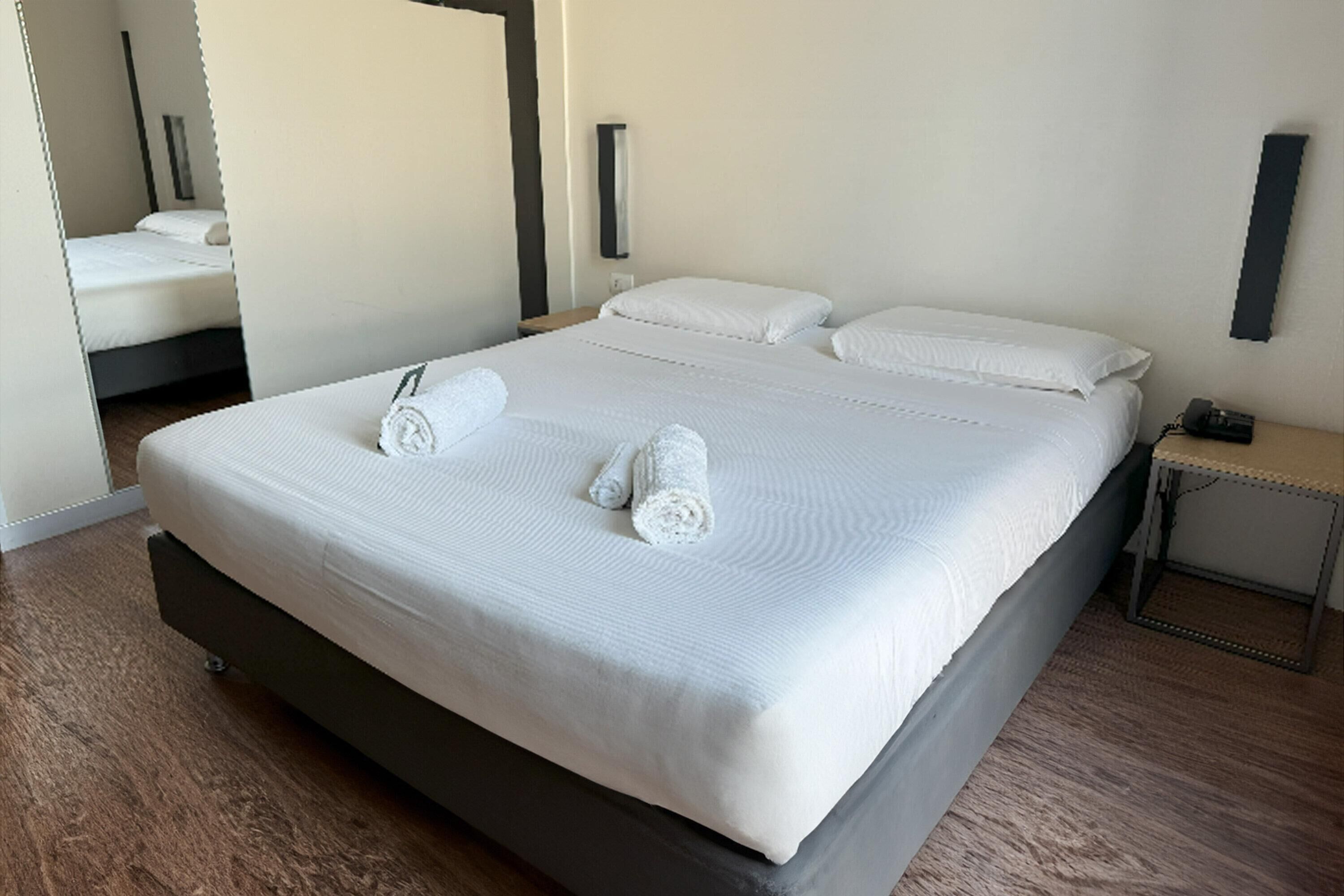 A neatly made bed with white linens and two rolled towels, a bedside table with a lamp, and a wooden floor.