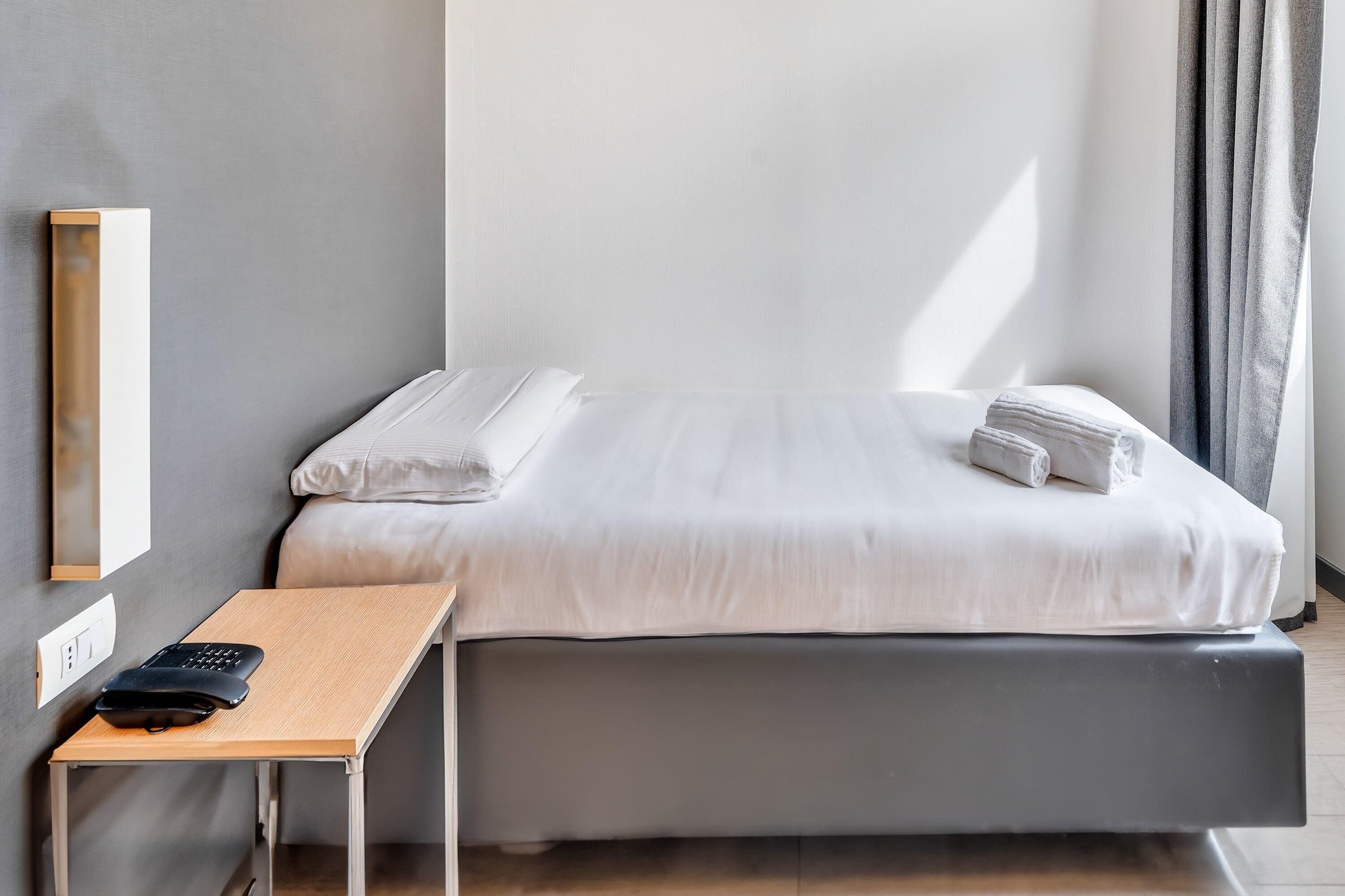 Ein ordentlich bezogenes Bett mit weißen Leinentüchern, ein kleiner Holztisch mit einem schwarzen Gegenstand darauf und ein Lichtschalter an der Wand.