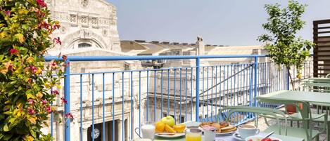 A balcony with a table set for breakfast, including bread, fruit, and beverages, overlooking a historic building with intricate architecture.