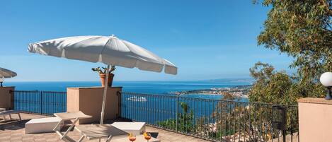 Una terraza con sombrilla blanca, sillones de descanso y una mesa con bebidas, con vistas a la costa.