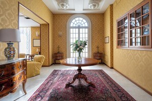 A room with a large arched window, a round wooden table with a vase of flowers, a yellow armchair, and a decorative cabinet. - Excelsior Palace Hotel (Taormina)