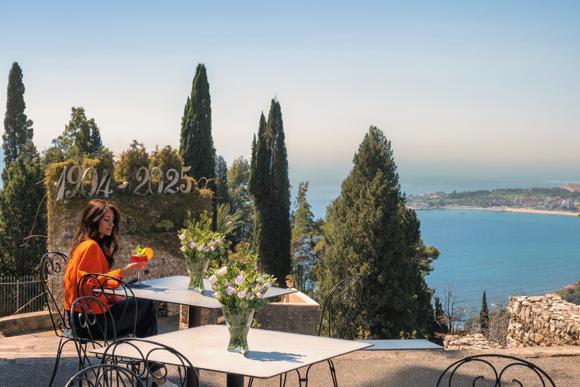 A table with a vase of flowers, a person sitting, and a view of the sea and trees.