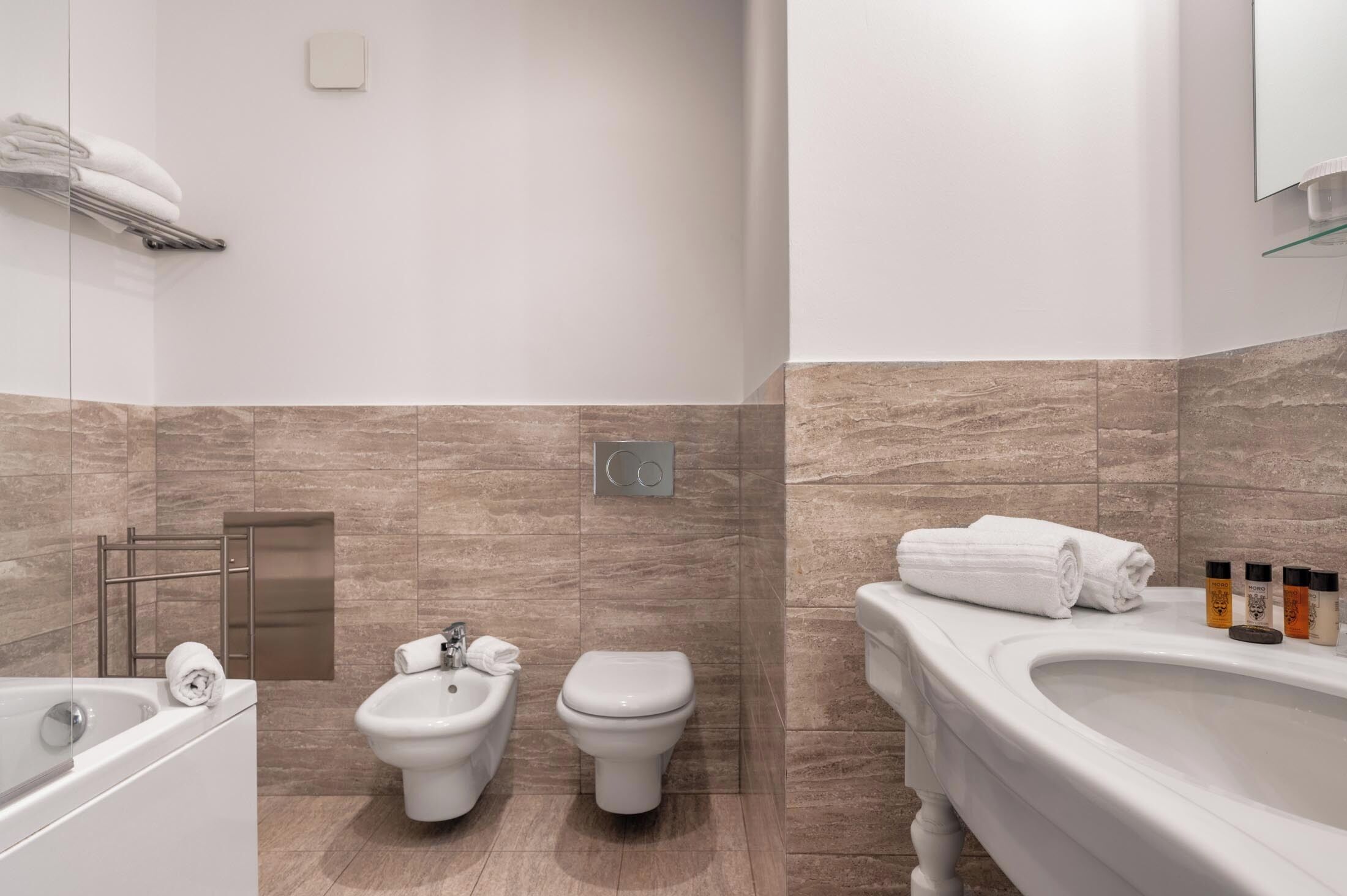 A modern bathroom with a bathtub, bidet, and toilet. There are towels on a rack and a glass shelf with toiletries.