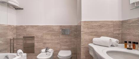 Een moderne badkamer met een badkuip, bidet en toilet. Handdoeken liggen op een rek en een glazen plank met toiletartikelen.