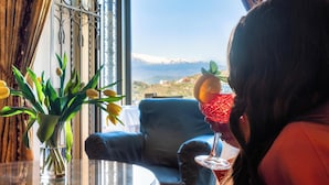 Una habitación con vistas a un paisaje montañoso, una mesa de cristal con un jarrón de flores amarillas y una persona sosteniendo una copa de cóctel.