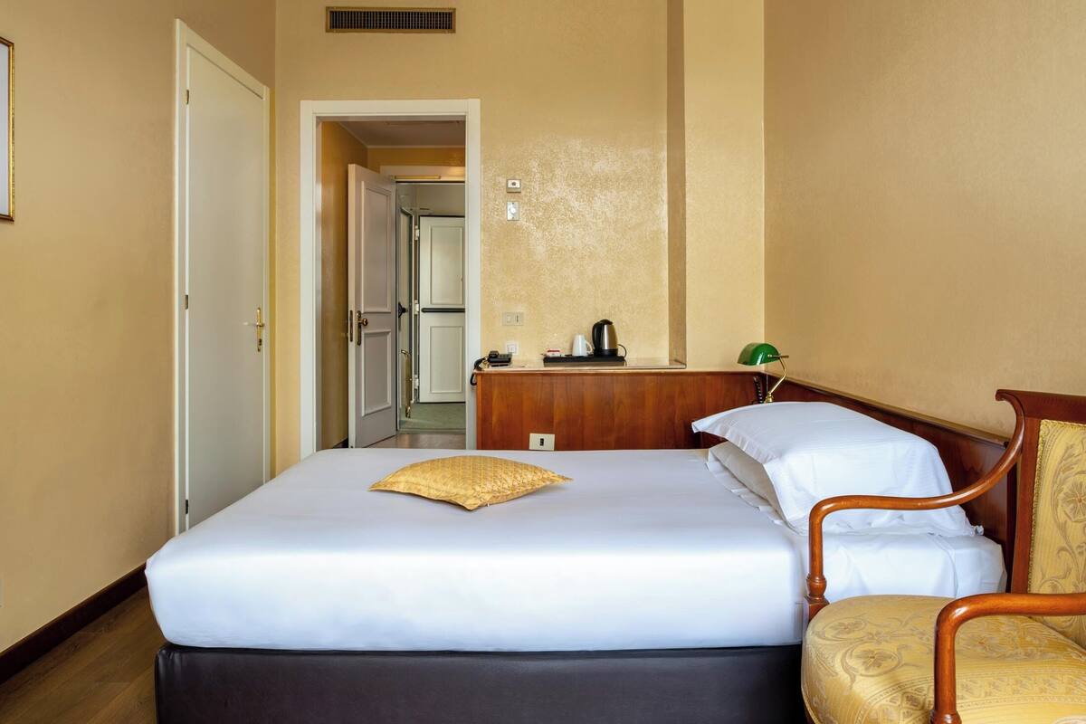 Una habitación de hotel con cama, una silla, una mesita de noche de madera y una puerta que da a otra habitación.