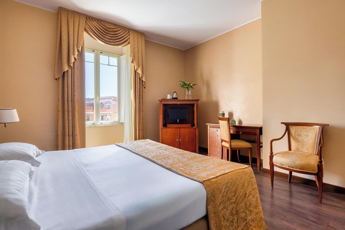 Una habitación de hotel con una cama grande, un escritorio con silla, televisión y una ventana con cortinas.