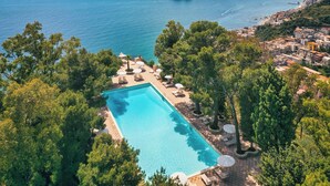 Vista aérea de un resort con piscina rodeada de sillones de descanso y sombrillas, con vistas a una zona costera con edificios y embarcaciones.