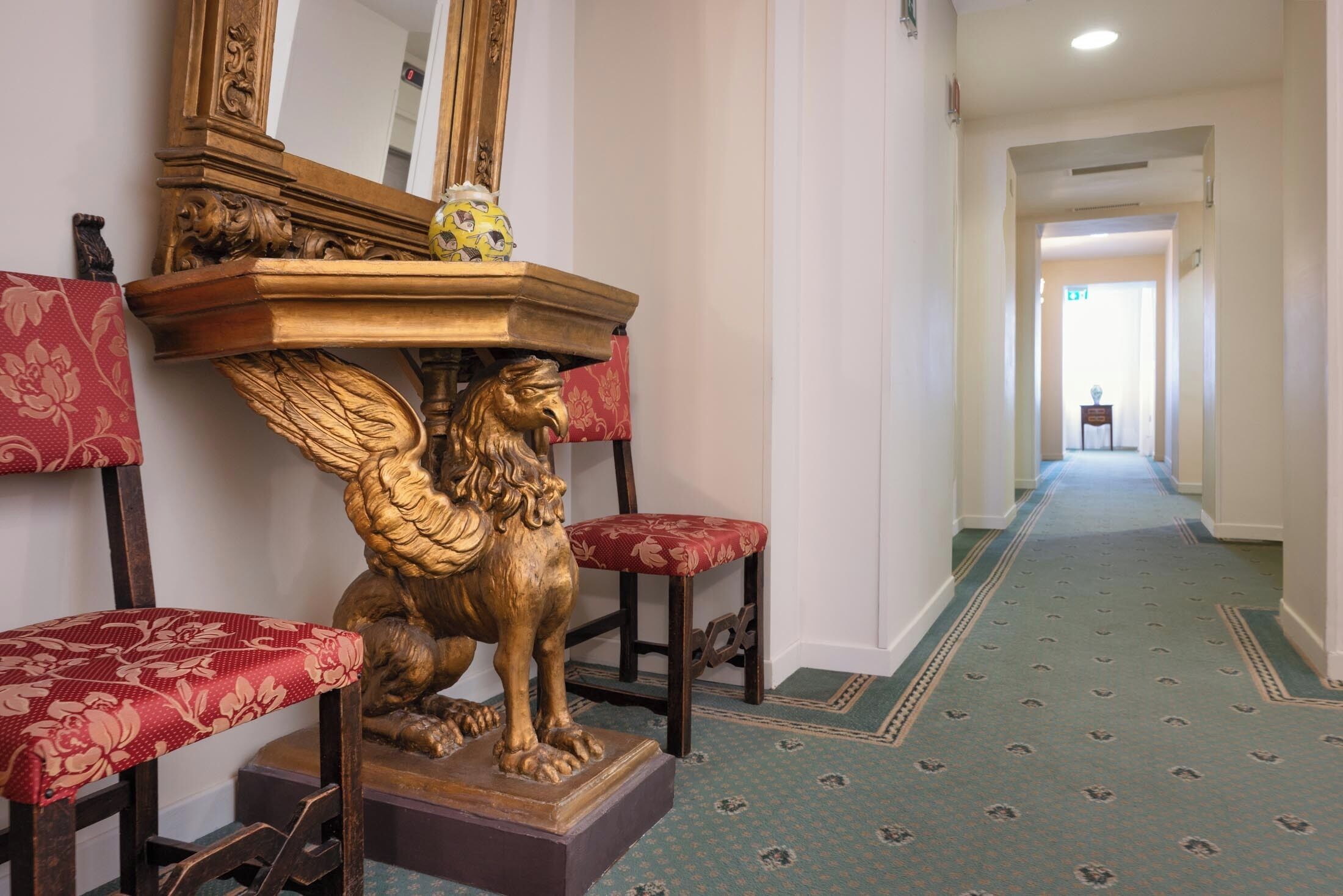 Un couloir orné d’une statue de lion doré, posée sur un piédestal, et flanqué de deux fauteuils à l’assise recouverte d’un tissu floral rouge.