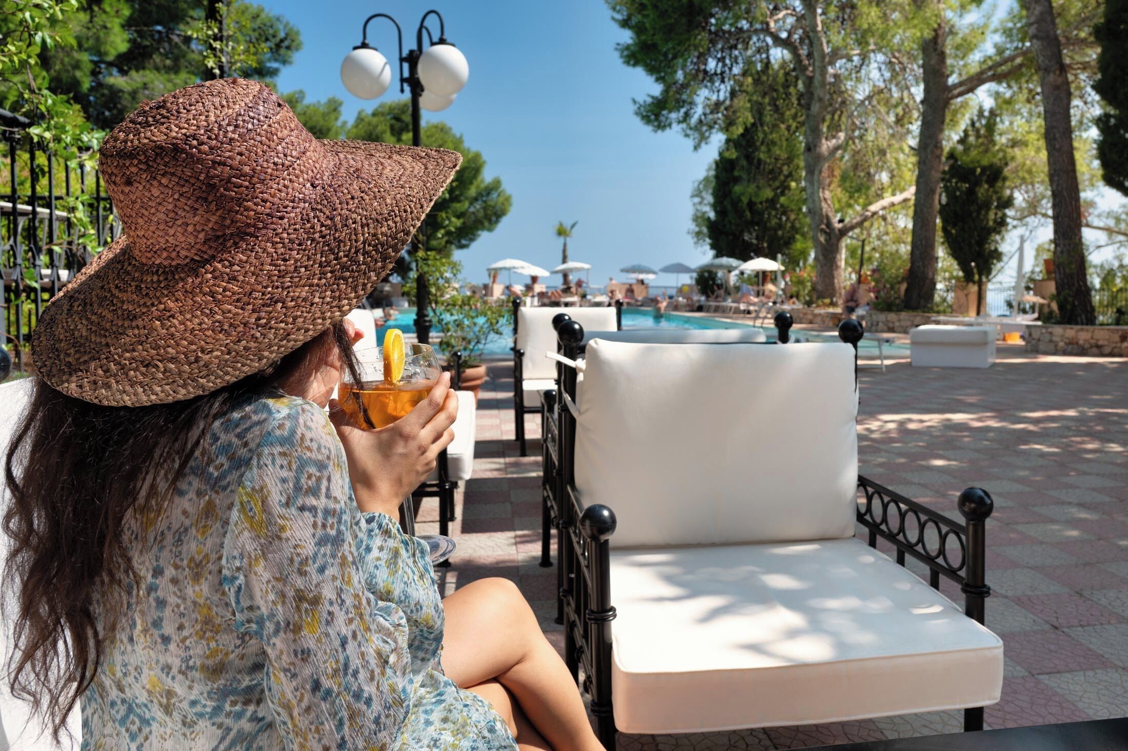 A person sitting on a white cushioned chair with a large woven hat, holding a drink, in a sunny outdoor area with a pool and umbrellas in the background.