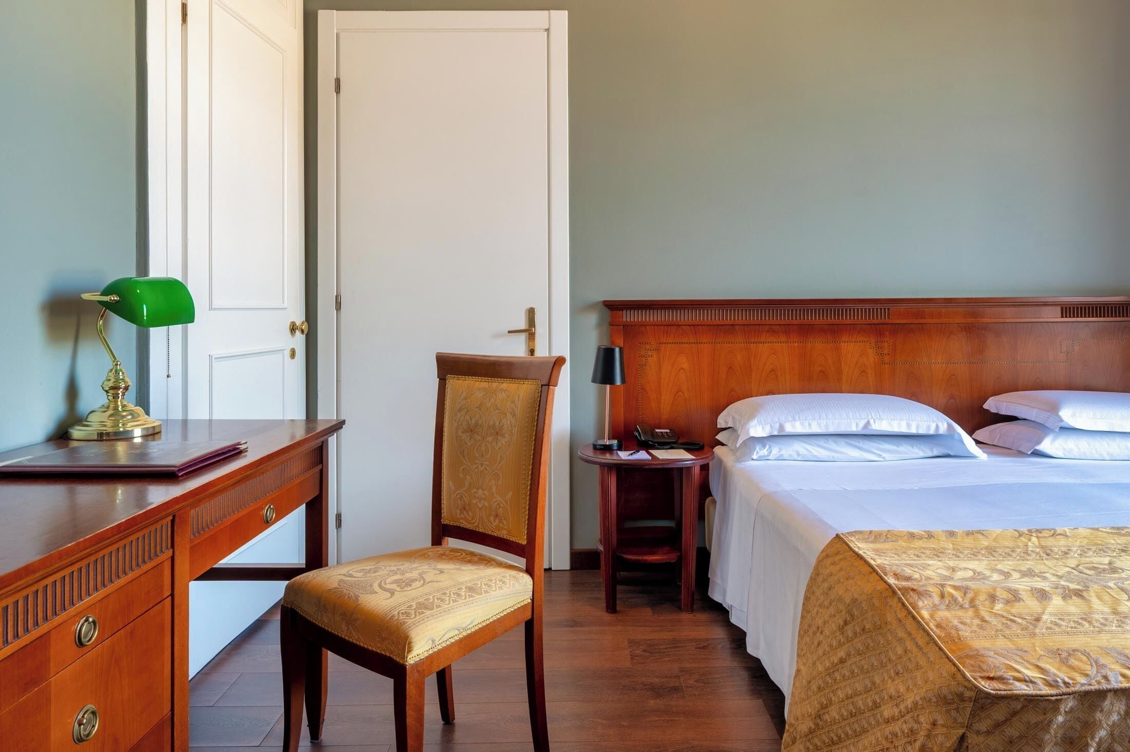 Une chambre d’hôtel avec un bureau en bois, une lampe de bureau verte, une chaise, une table de chevet avec une lampe, un lit avec du linge de lit blanc et un plaid à motifs.