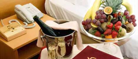 A hotel room with a bed, a bedside table with a phone, a champagne bottle, a wine glass, and a fruit basket.