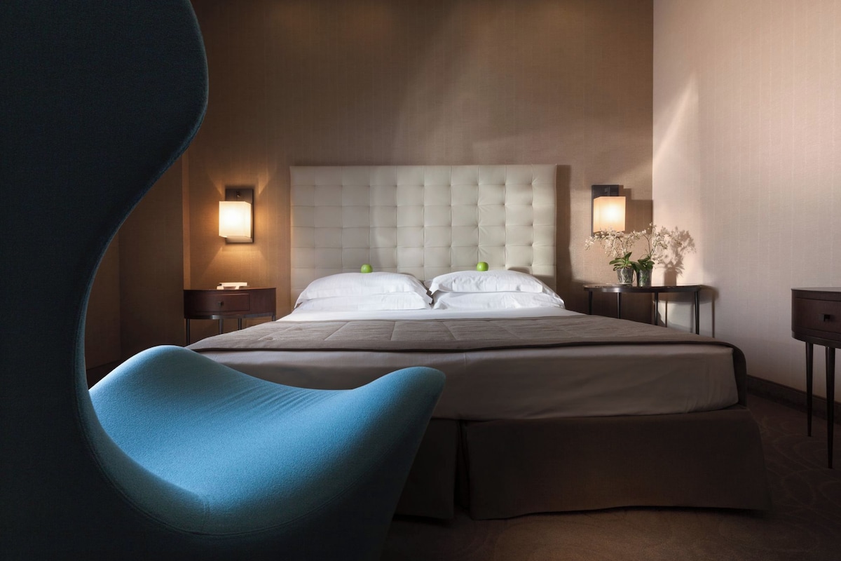 A hotel room with a large bed, two bedside tables, a nightstand, a blue chair, and a wall-mounted lamp.