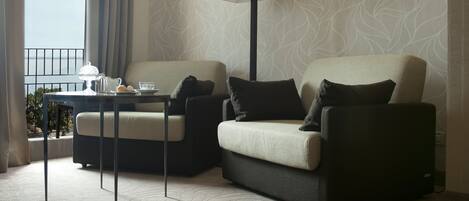 A modern hotel room with two sofas, a table with a lamp, and a view of the sea.