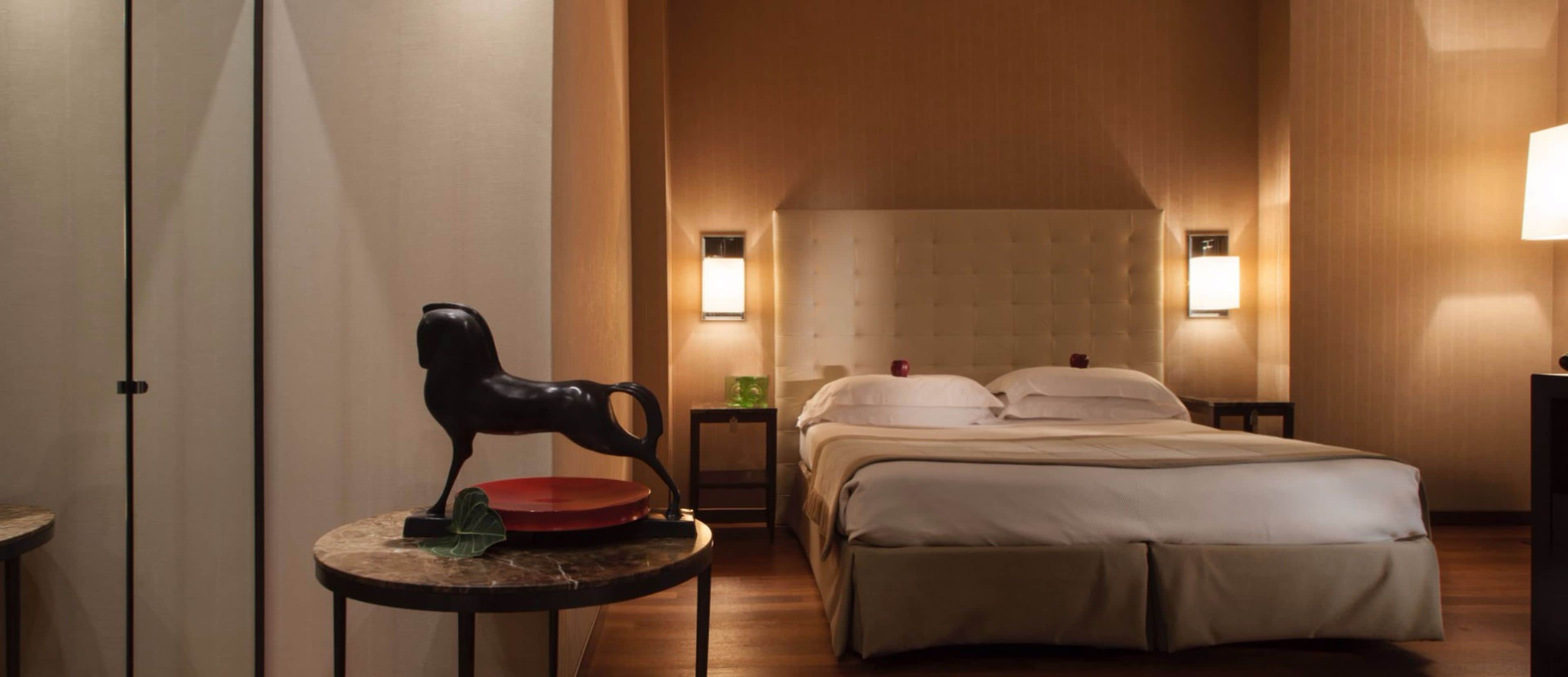 Hotellihuone, jossa on sänky, yöpöydät, lamppu ja koristeellinen hevospatsas.
