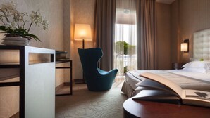 Habitación de hotel con una cama grande, una silla azul, una mesita de noche con lámpara y vistas al exterior a través de la ventana.