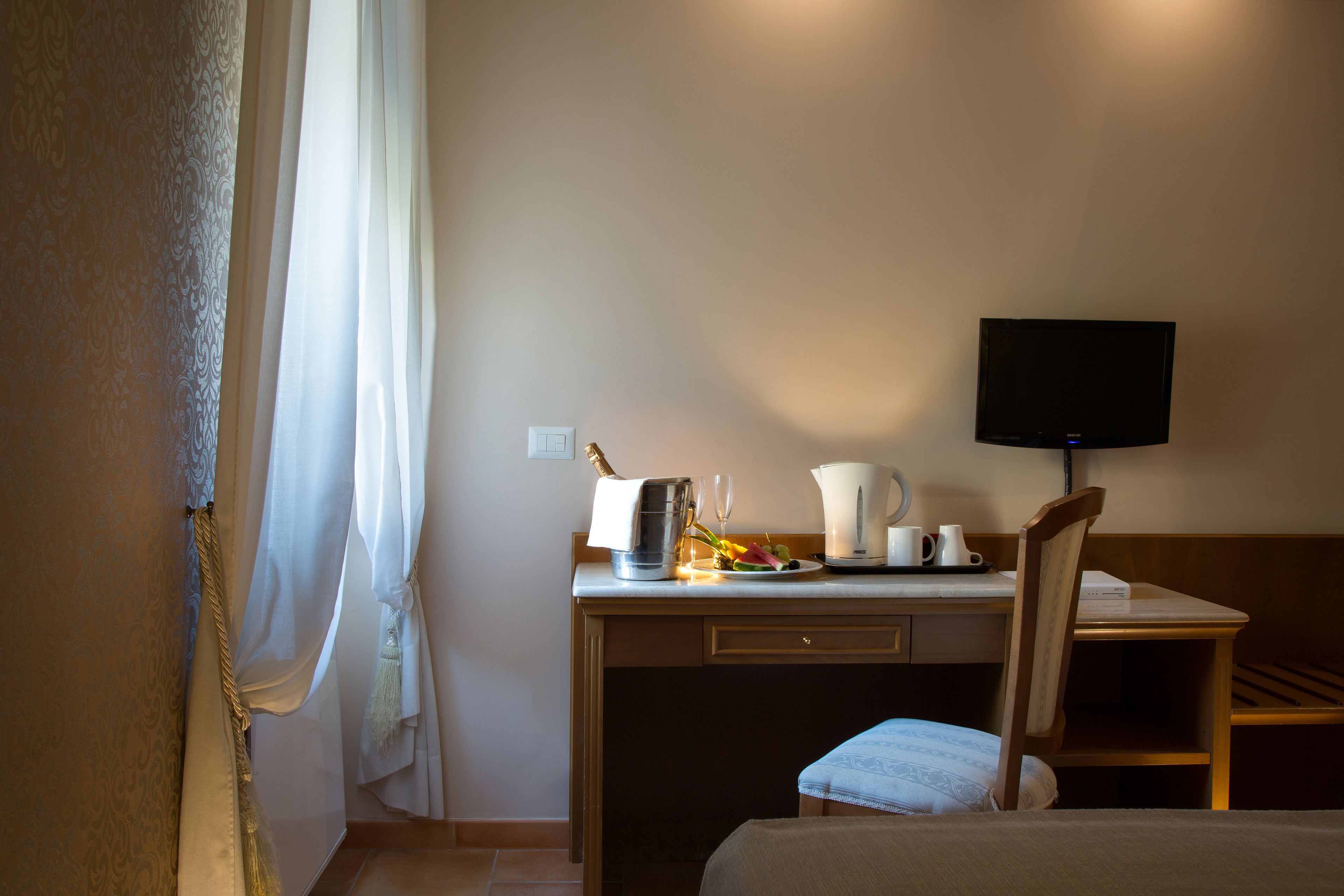 Ein Hotelzimmer mit Bett, Schreibtisch mit Fernseher, Stuhl und kleiner Tisch mit Wasserkocher, Kaffeemaschine und einer Platte mit Blumen.
