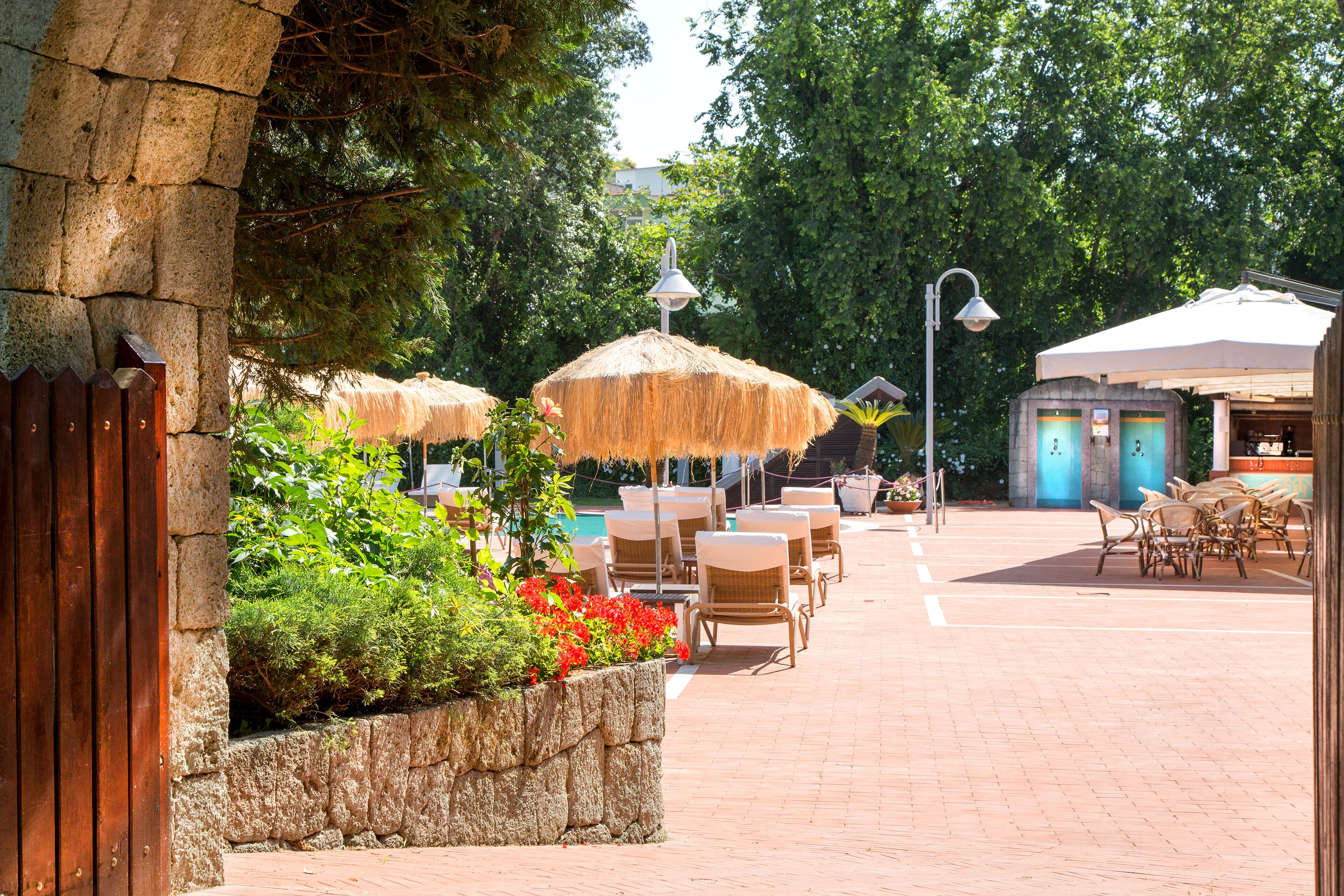 An outdoor seating area with wicker chairs and thatched umbrellas, a stone wall with greenery, and a building with a blue door in the background.