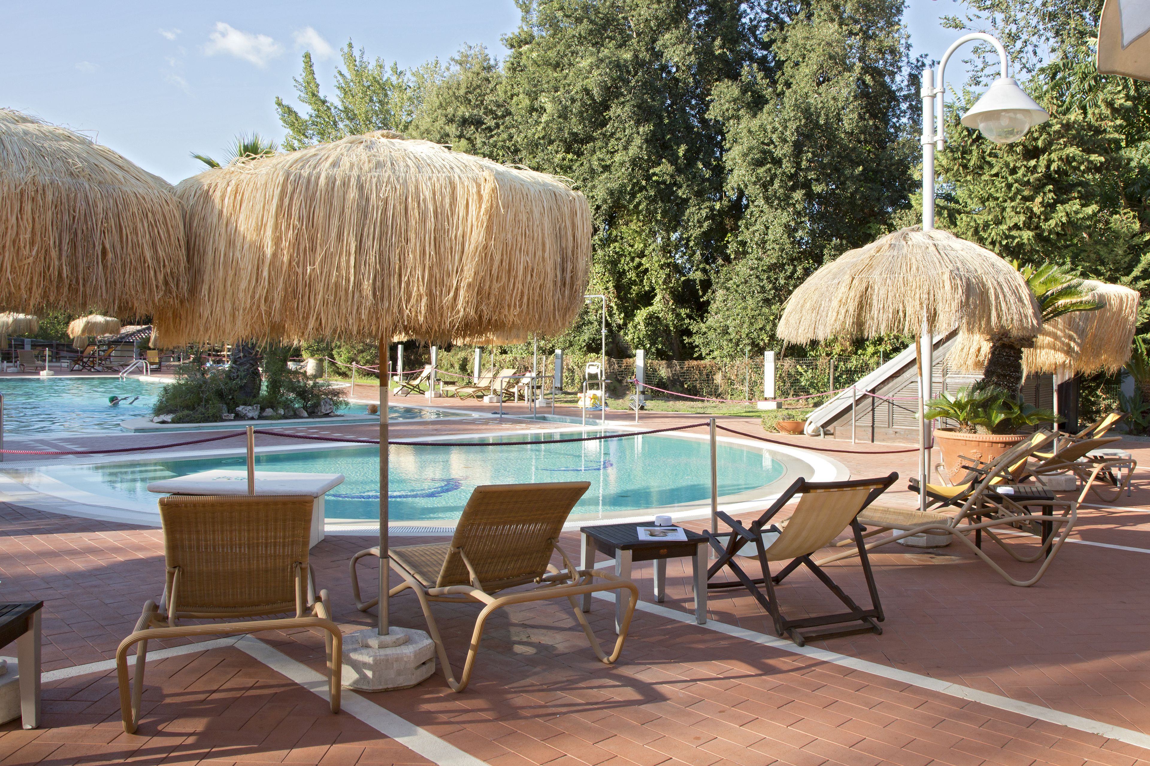 A poolside area with lounge chairs and thatched umbrellas, surrounded by trees and a clear sky.