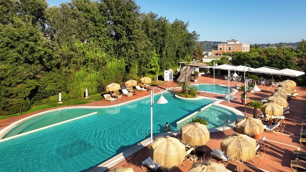 A resort pool area with lounge chairs and umbrellas, surrounded by trees and a building in the background. - Montespina Park Hotel (Naples)