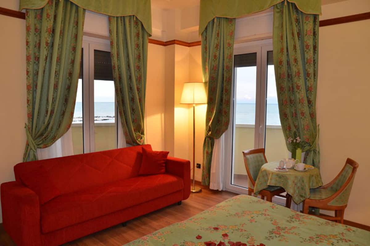 Habitación con sofá rojo, una pequeña mesa con sillas y vista al mar a través de amplias ventanas con cortinas florales.