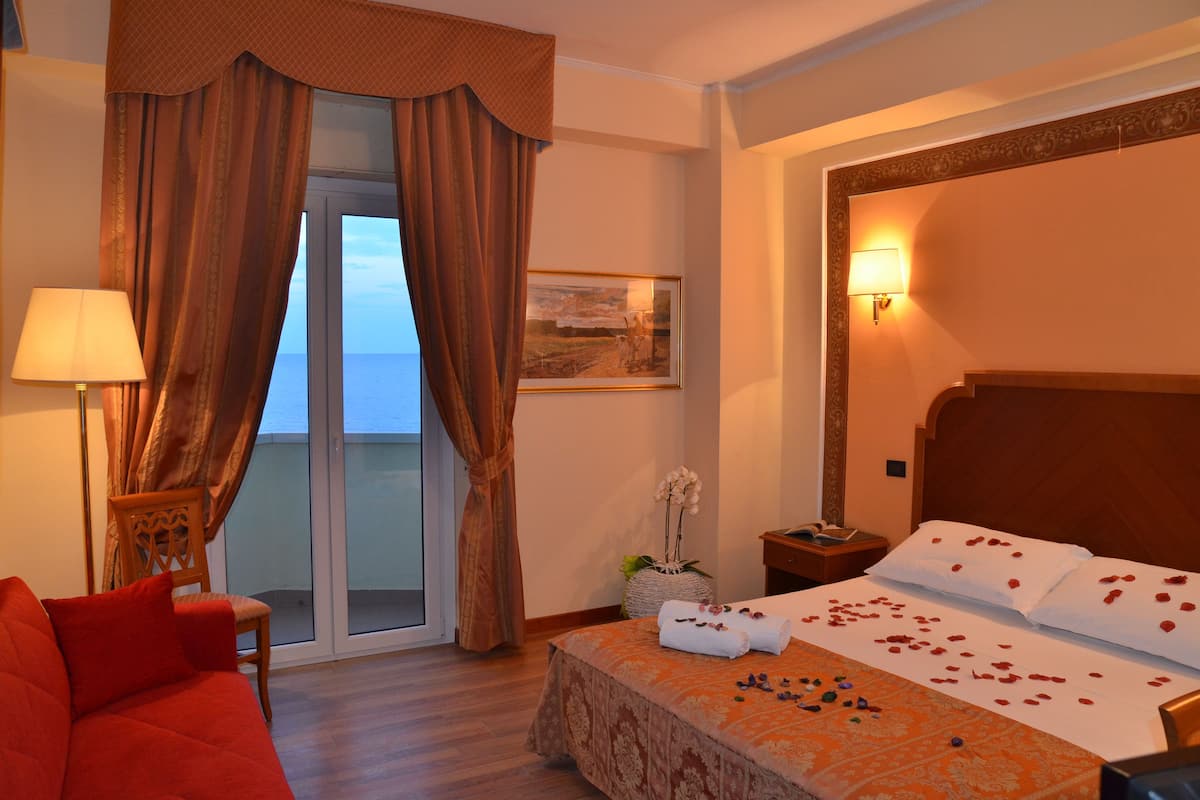 Una habitación de hotel con una cama, un sofá rojo, una mesita de noche, una lámpara y vistas al mar a través de una puerta corredera.
