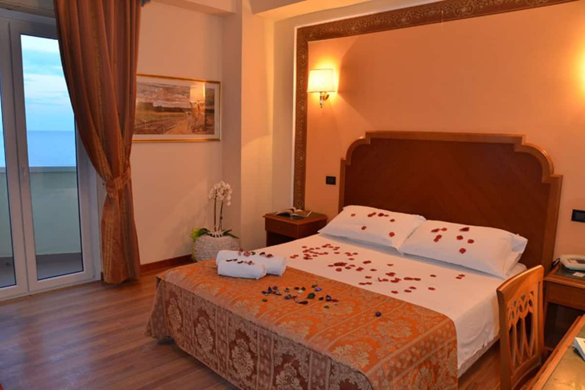 Habitación de hotel con cama, mesita de noche, lámpara, ventana con cortinas y vista al mar.