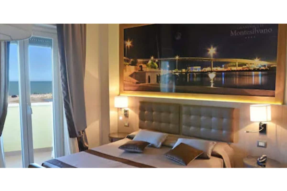 Habitación de hotel con cama, mesita de noche, lámpara y un amplio ventanal con vista a la playa.
