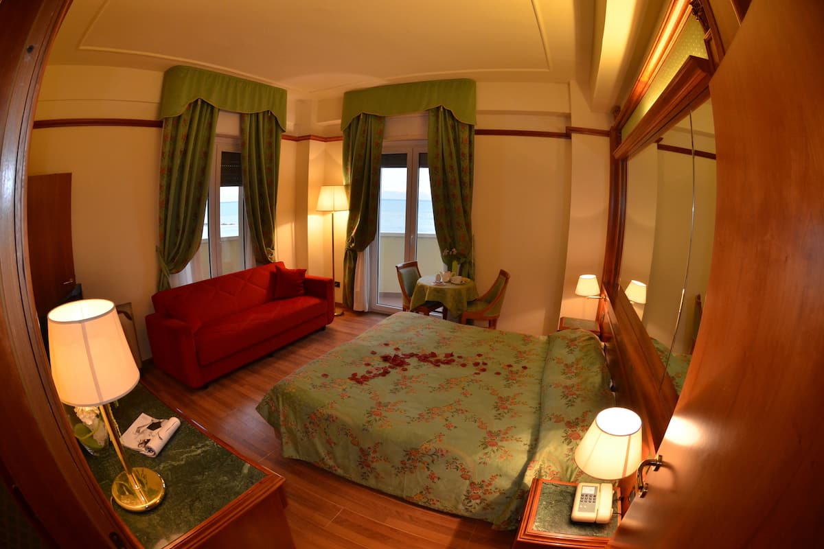 Una habitación de hotel con una cama, un sofá rojo, una mesita con una lámpara y un espejo.