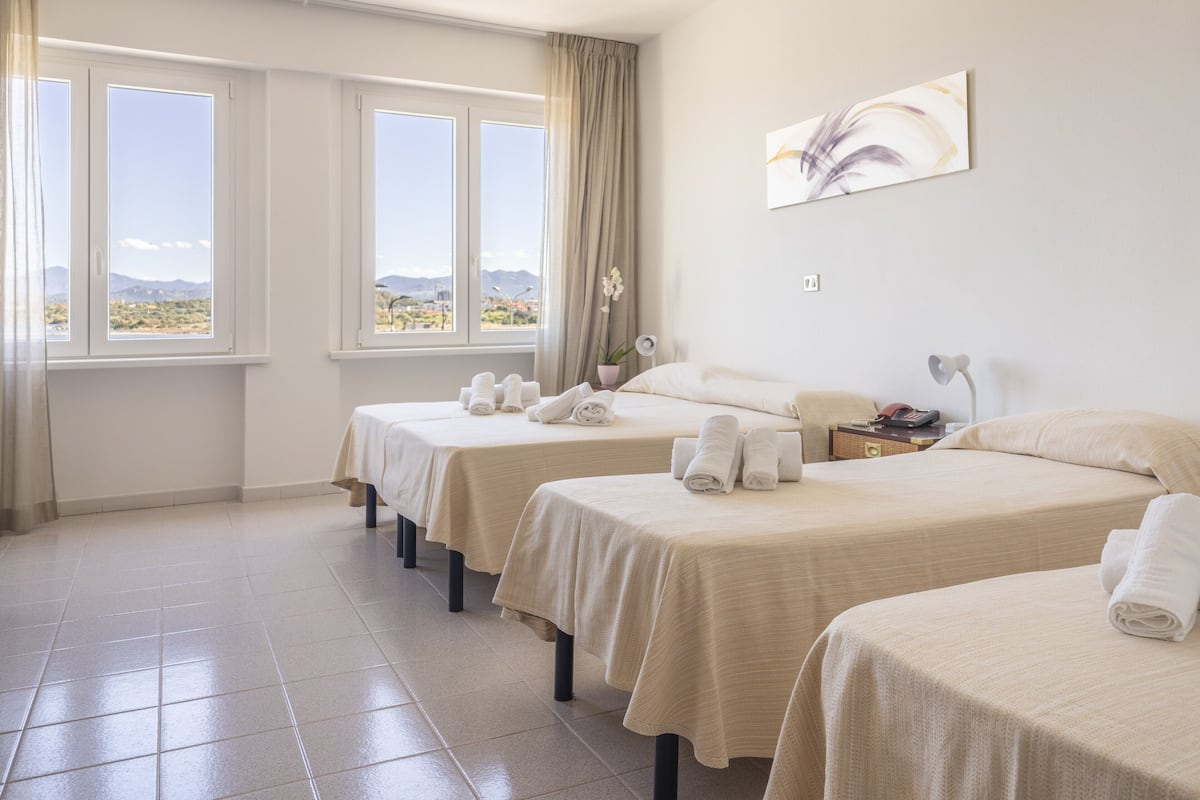 Une chambre d’hôtel avec trois lits, une grande fenêtre donnant sur le désert, un tableau au mur et une table de chevet avec une lampe.