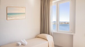 A single bed with a beige bedspread and a rolled towel, a window with a view of a body of water and distant land, and a framed picture on the wall. - De Plam (Olbia)