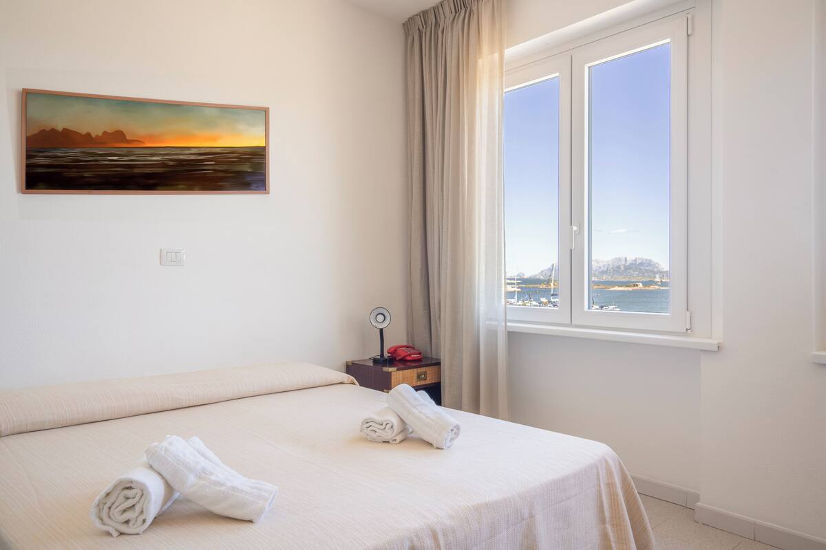 Une chambre d’hôtel avec un lit, un tableau représentant un coucher de soleil, une fenêtre donnant sur la mer et une table de chevet avec une lampe et un téléphone.