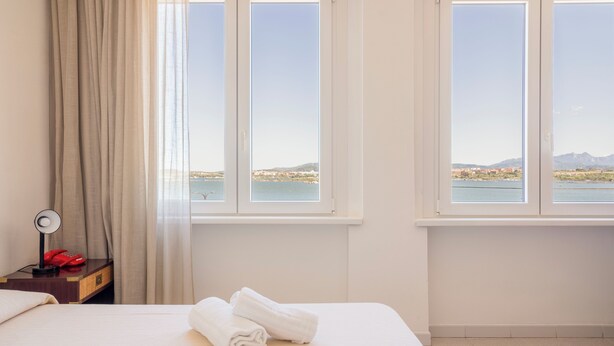 A hotel room with a bed, a nightstand with a lamp and a red telephone, and two windows offering a view of a body of water and distant land. - De Plam (Olbia)