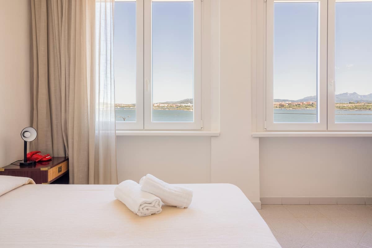Une chambre d’hôtel avec un lit, une table de chevet dotée d’une lampe et d’un téléphone rouge, et deux fenêtres donnant sur une étendue d’eau et un paysage lointain.