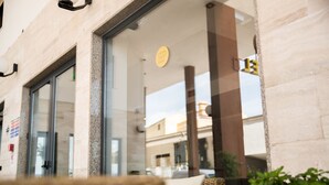 A modern building with large glass windows, a visible sign with a logo, and a reflection of the sky and buildings in the glass. - De Plam (Olbia)