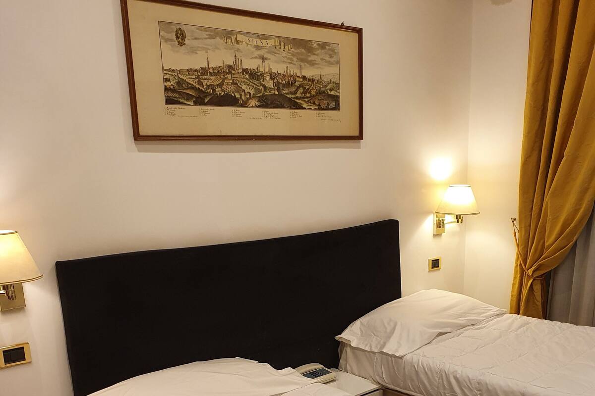 Une chambre d’hôtel avec deux lits, une table de chevet, une lampe, un tableau représentant un paysage urbain encadré et un rideau.