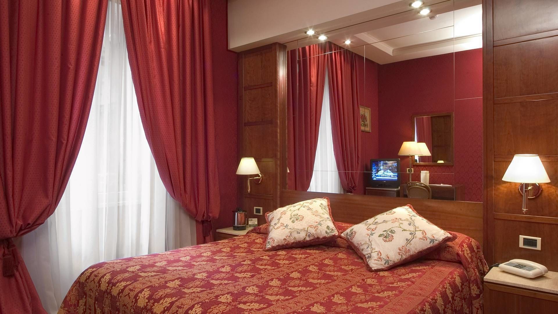 Ein Hotelzimmer mit einem großen Bett, roten Vorhängen, einem hölzernen Kopfteil, zwei Nachttischlampen, einem Fernseher und einem Telefon auf dem Nachttisch.