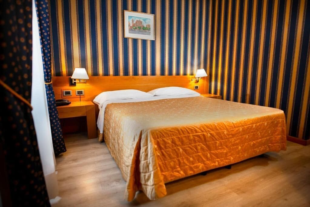 Habitación de hotel con una cama grande, dos mesitas de noche con lámparas, un cabecero de madera y papel tapiz a rayas.