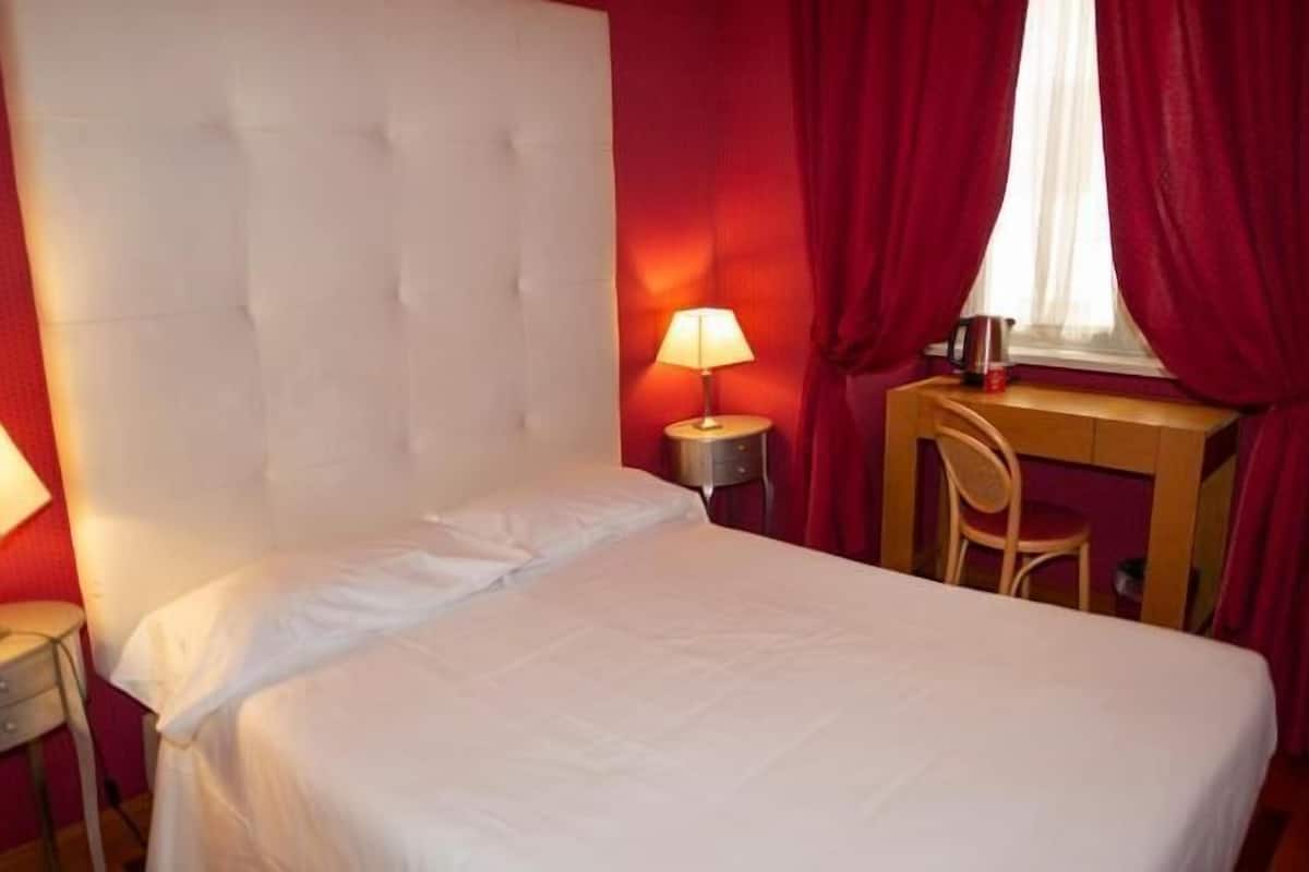 Une chambre d’hôtel avec un lit, un bureau, une chaise, une lampe et une fenêtre avec des rideaux.