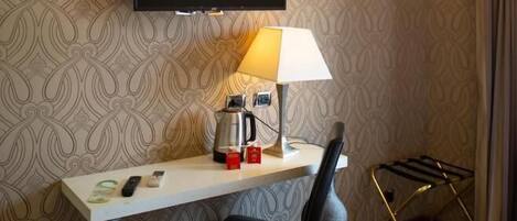 Habitación de hotel con escritorio, silla, televisión montada en la pared, lámpara, tetera y un pequeño cubo de basura.
