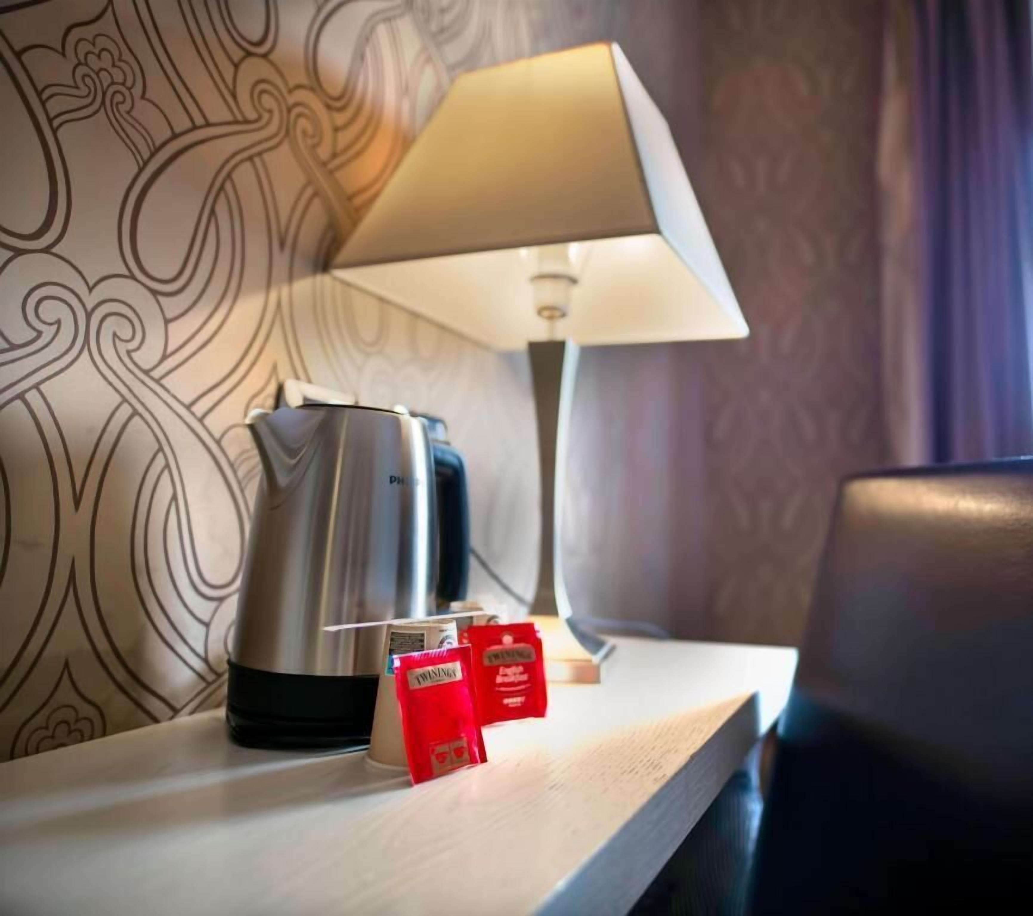 A hotel room with a coffee maker, a lamp, and two red packets on a wooden table.
