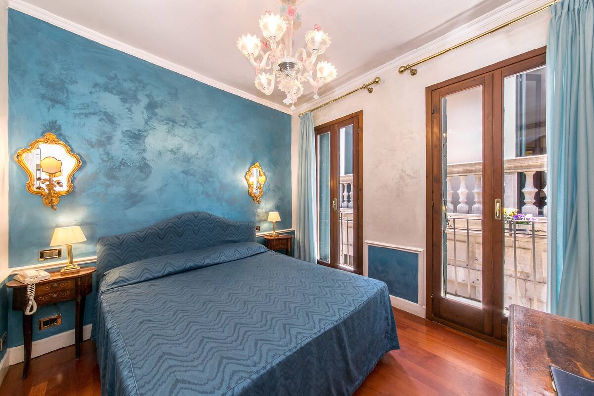 A bedroom with a large bed, two bedside tables with lamps, a chandelier, and a balcony with a view.
