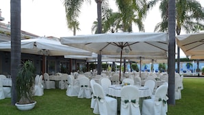 An outdoor dining area with white tables and chairs, covered by large umbrellas, surrounded by palm trees. - Hotel Orizzonte (Acireale)