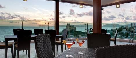 A restaurant with wicker furniture and a view of the sea and cityscape.
