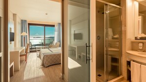 A modern hotel room with a bed, desk, and chair, a glass shower enclosure, and a view of the sea. - Smy Carlos V Alghero (Alghero)